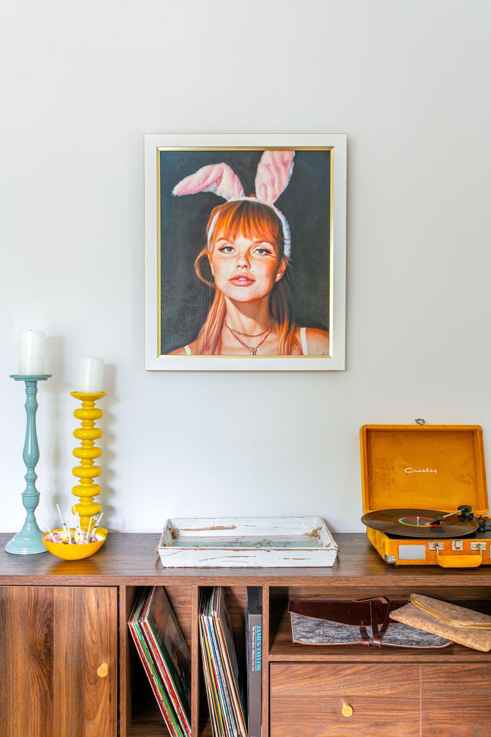 Retro wooden sideboard with vinyl records, yellow record player, candles, and a framed portrait painting of a woman with bunny ears.