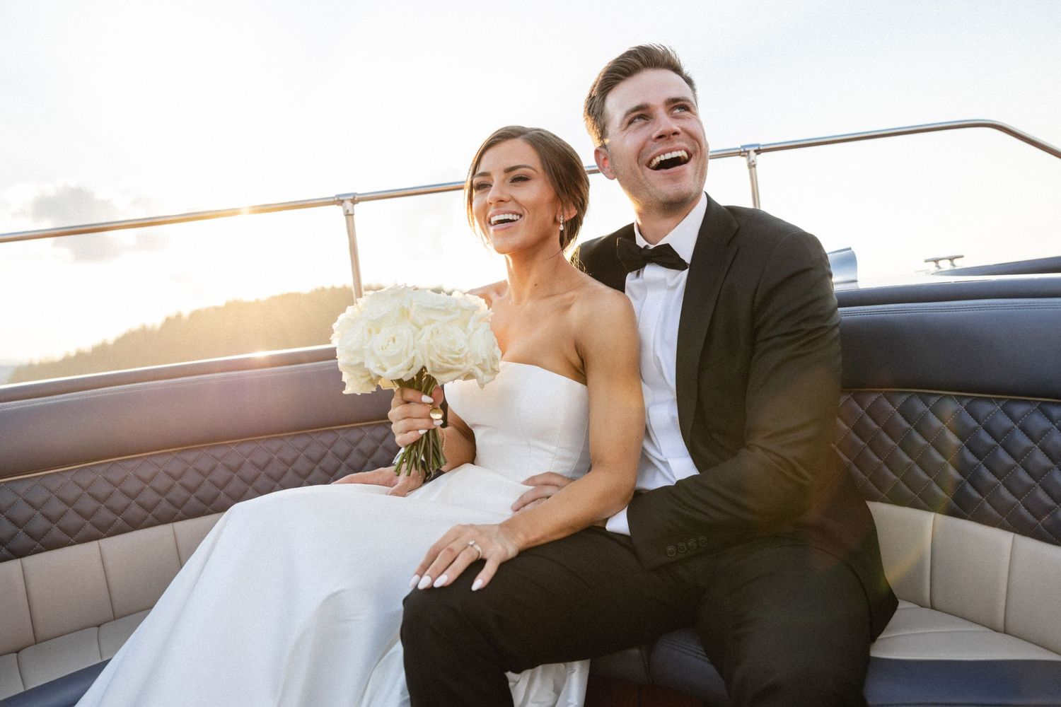 Bride and groom pose by waterfront hedge at sunset, she in strapless gown, he in black tuxedo.
