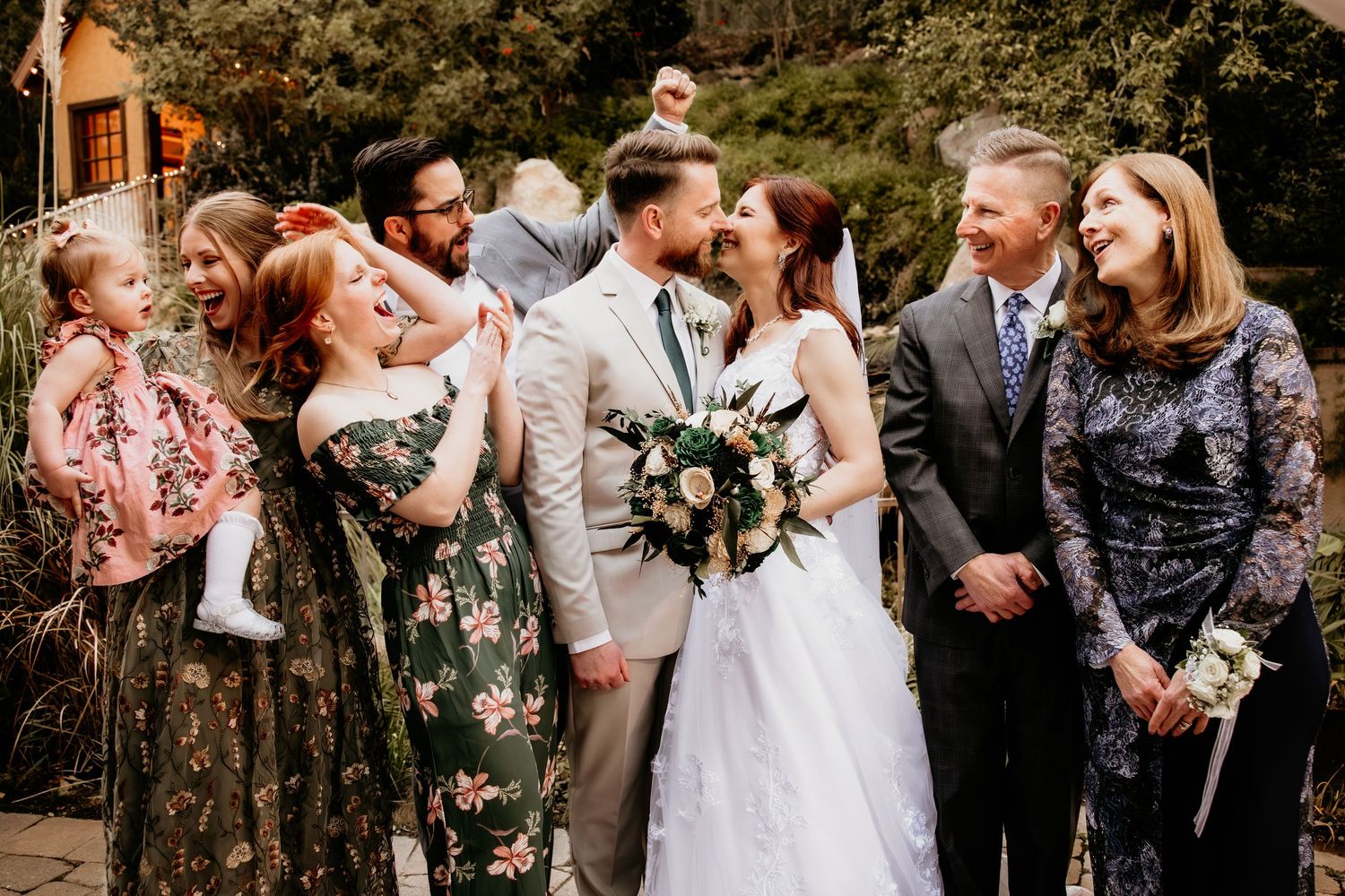 A newlywed couple kisses surrounded by smiling family members during an outdoor wedding celebration.