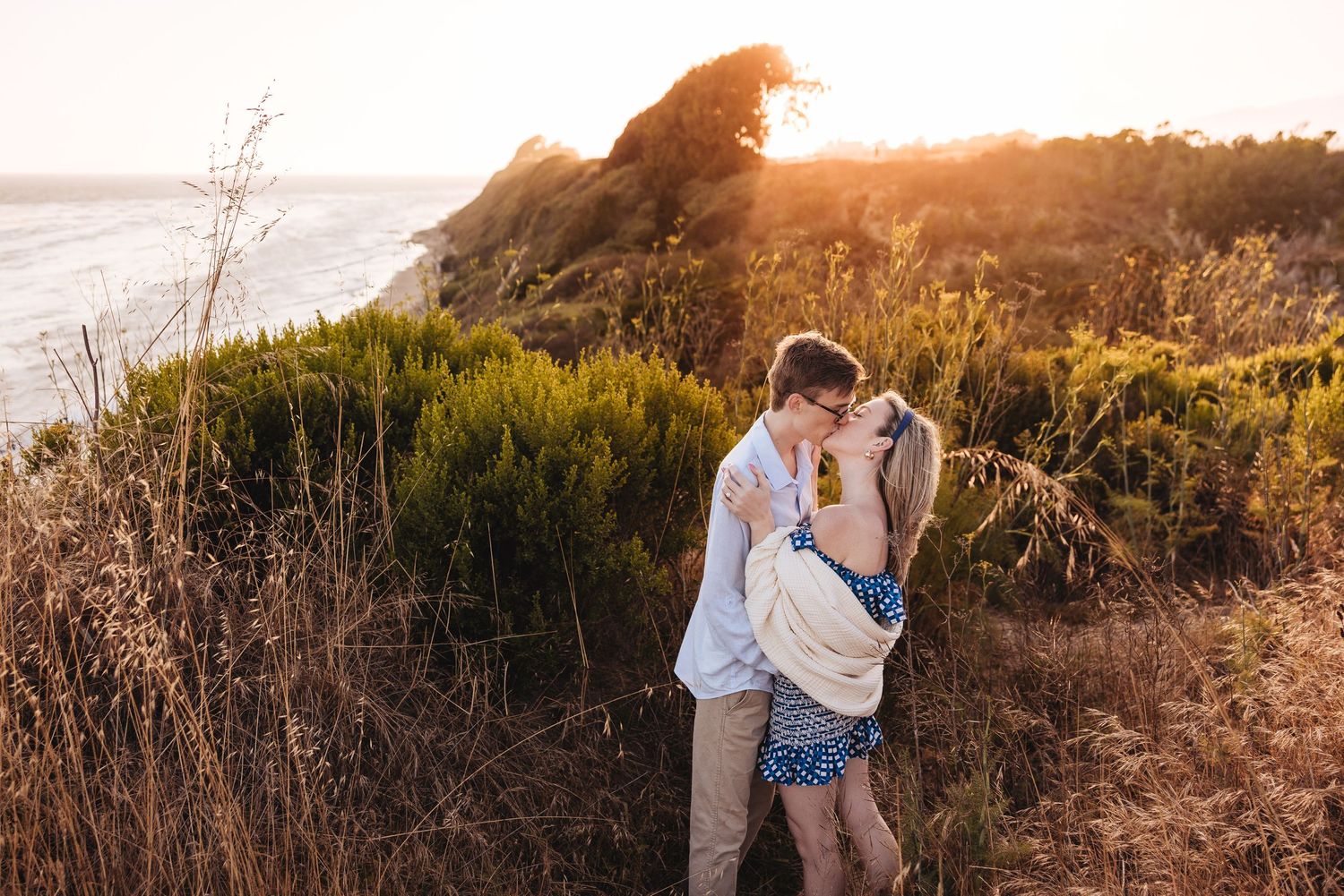 Couple sharing a romantic kiss on a coastal cliff at golden sunset surrounded by lush greenery and ocean views.