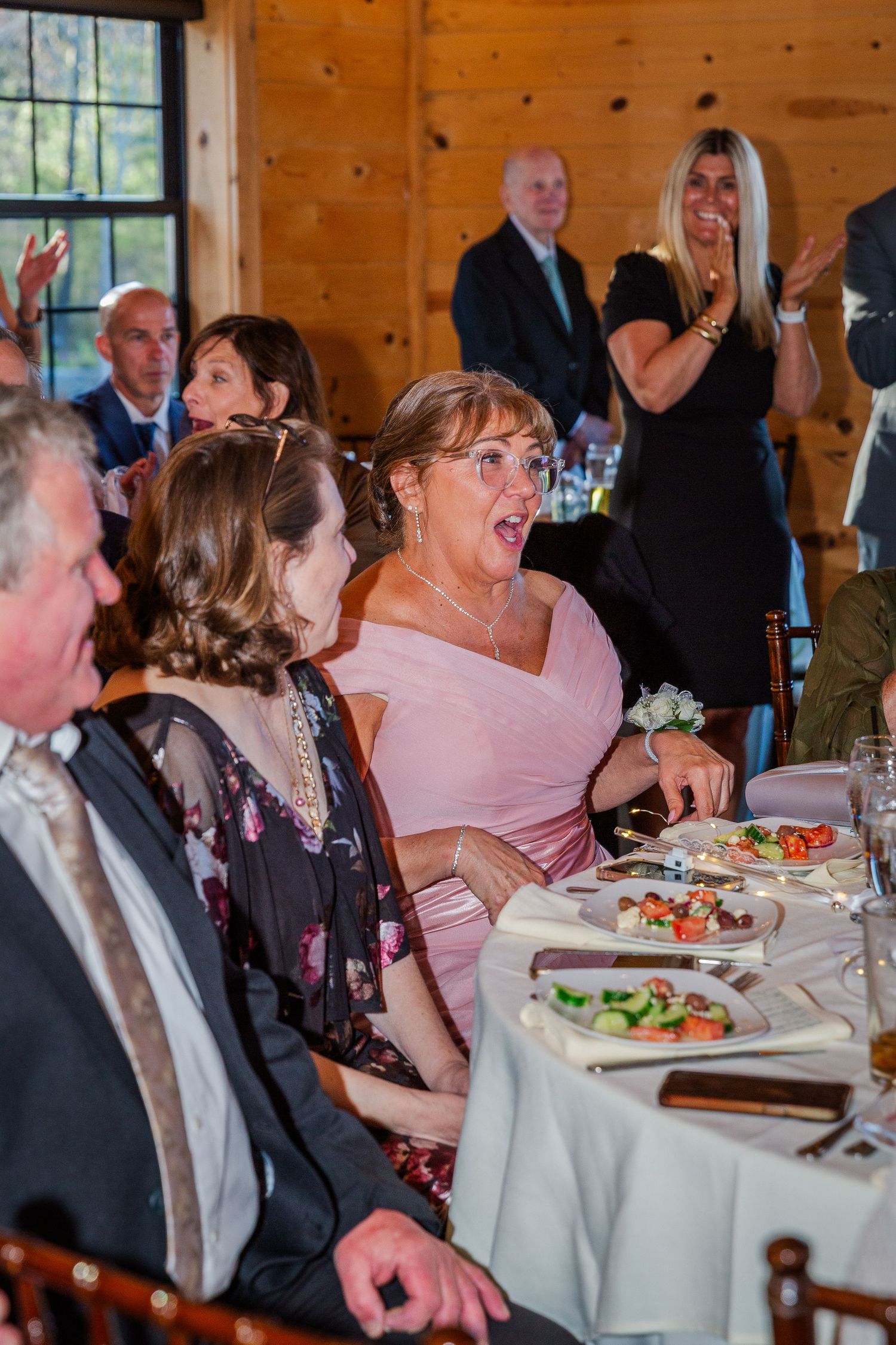 Wedding reception guests react with surprise and joy at a rustic wooden venue during a celebratory dinner.