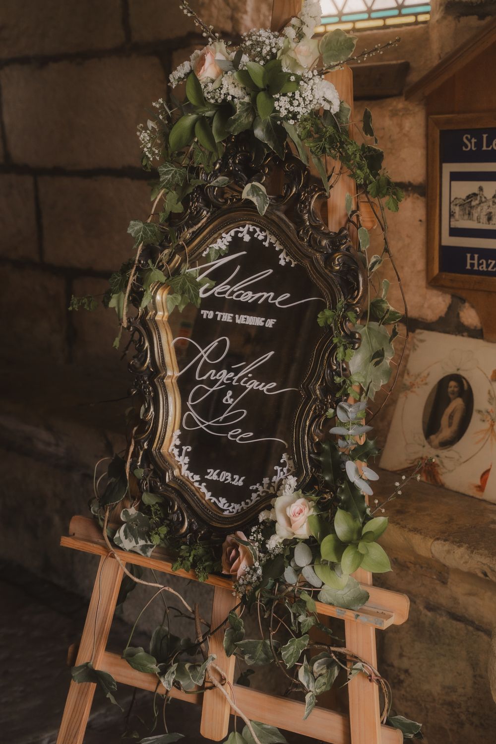 Elegant wedding welcome mirror sign with floral greenery on wooden easel inside a stone church.