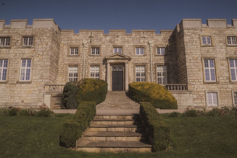Historic stone manor house with grand entrance steps, manicured hedges, and castellated roofline under a clear blue sky.