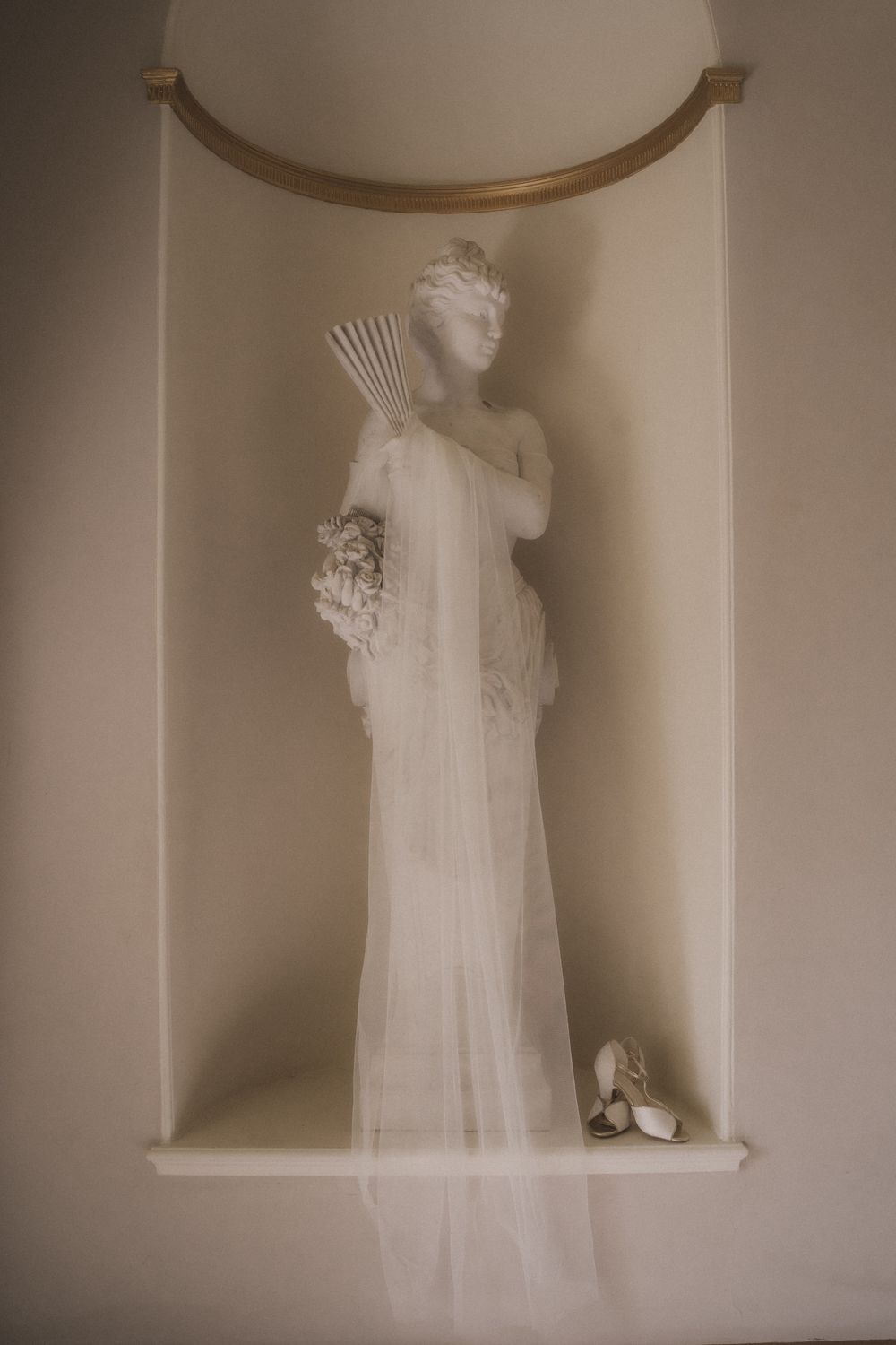 White classical statue of a goddess in a wall niche, draped with a bridal veil, holding flowers and a fan.