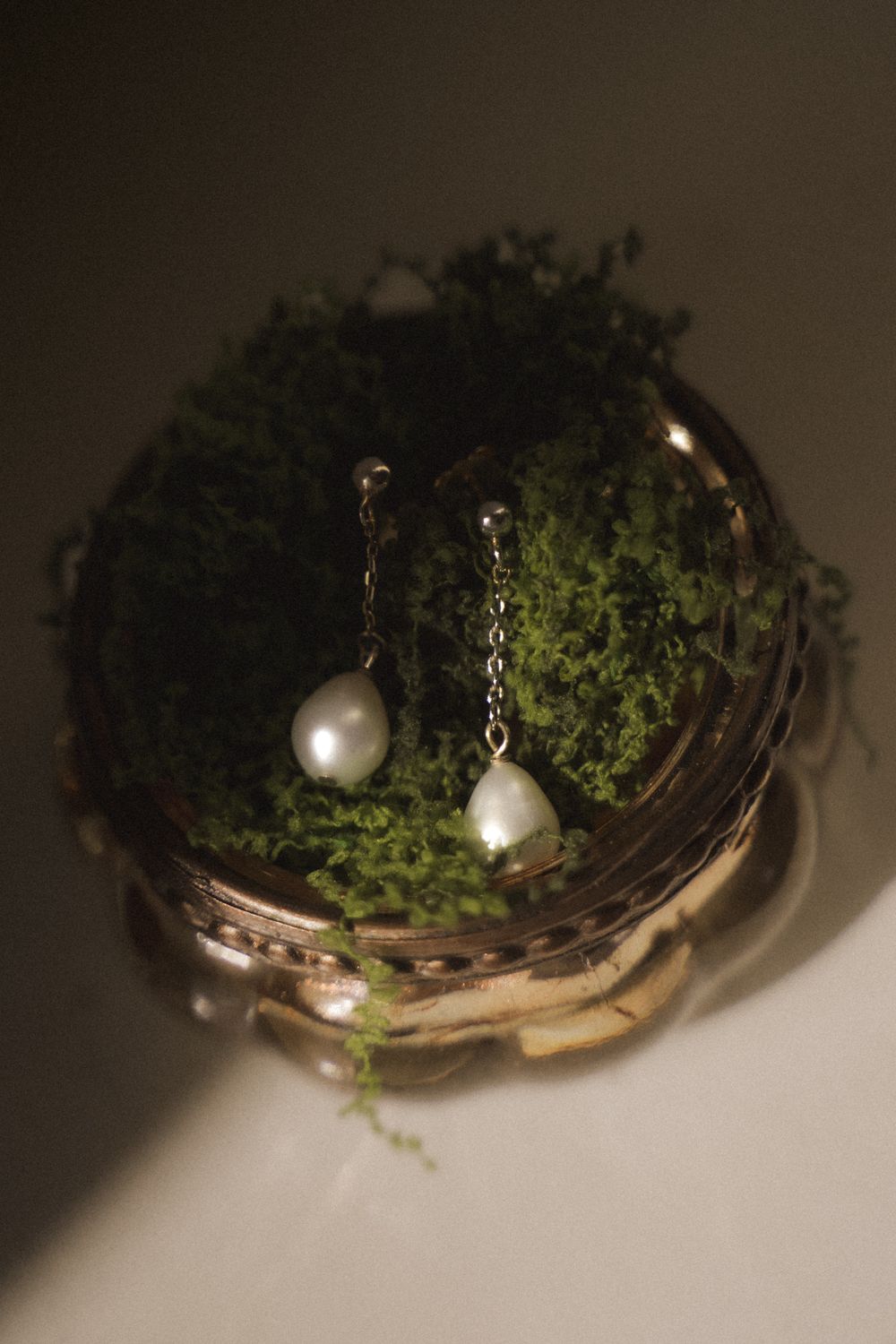 Elegant pearl drop earrings with delicate chain details resting on green moss inside a vintage gold jewelry box.