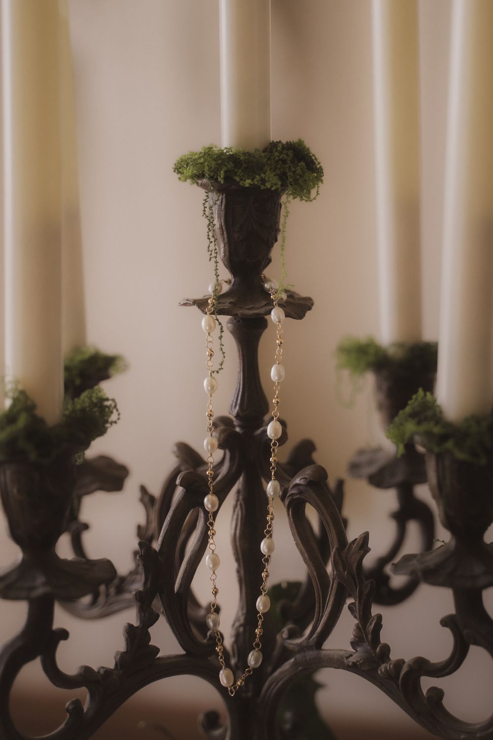 Ornate dark metal candelabra with white taper candles, green moss accents, and hanging pearl jewelry chain centerpiece.