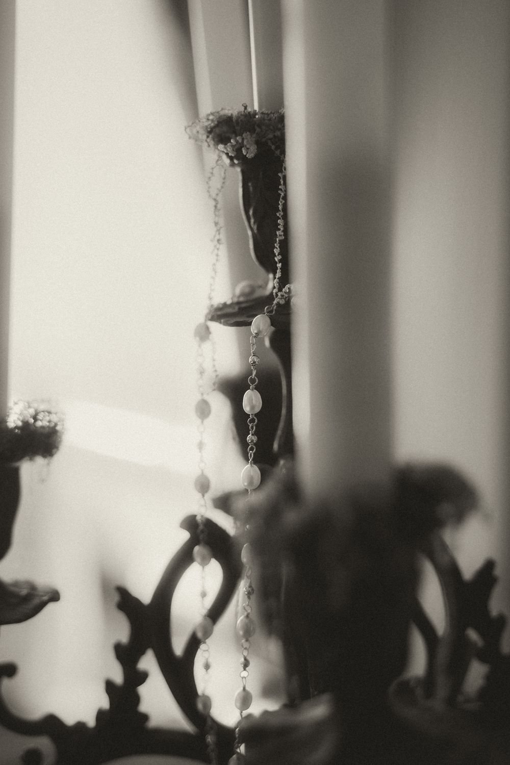 Black and white photo of elegant jewelry draped on a vintage candelabra near a bright window.