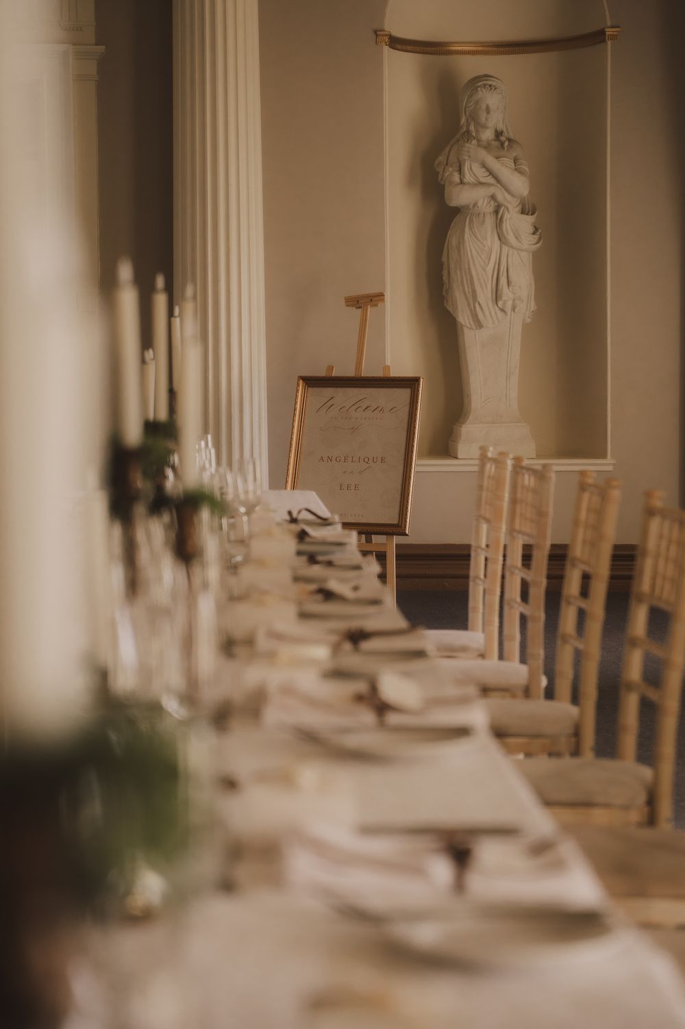 Elegant wedding reception hall with long dining table, candles, white statue, and a welcome sign on an easel.