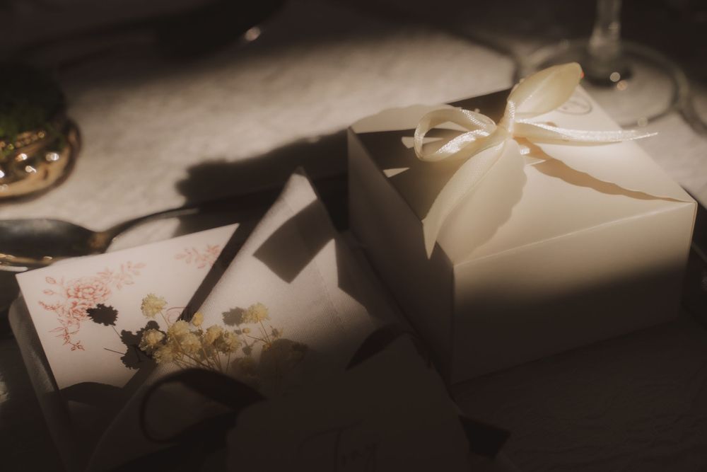 Elegant white gift box with cream ribbon bow beside a floral card and dried flowers on a softly lit table.