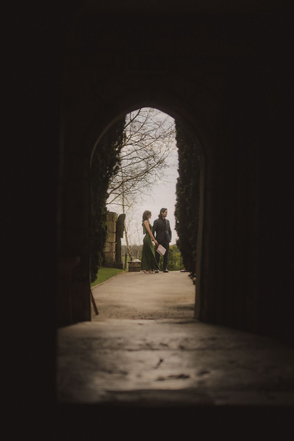 Two people stand framed by a dark stone archway tunnel, silhouetted against soft natural light beyond.