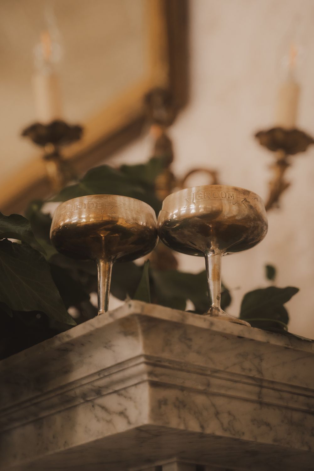 Two vintage gold champagne coupe glasses on a marble mantle with candles and greenery in warm ambient light.