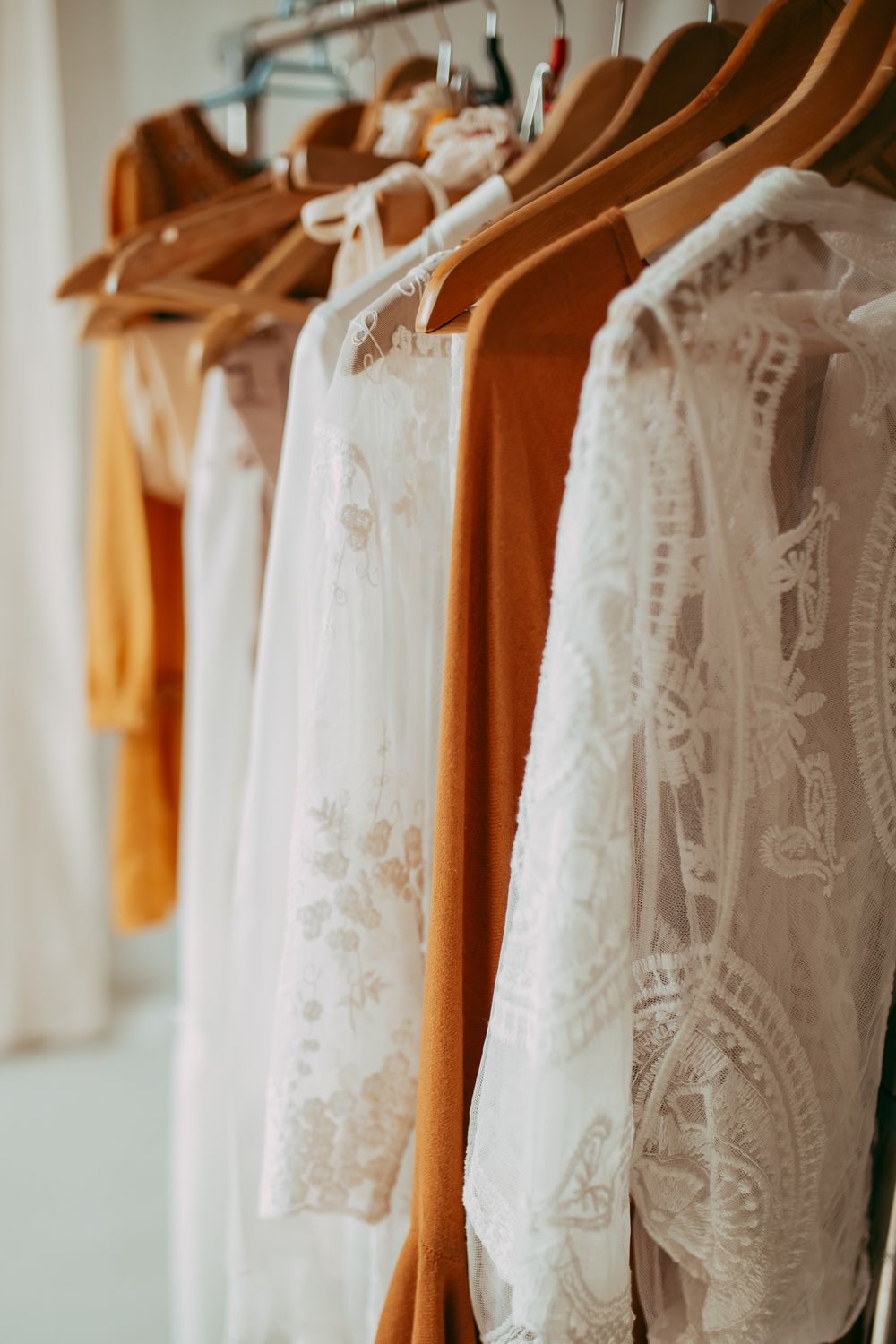 White lace and burnt orange dresses hanging on wooden hangers on a clothing rack.