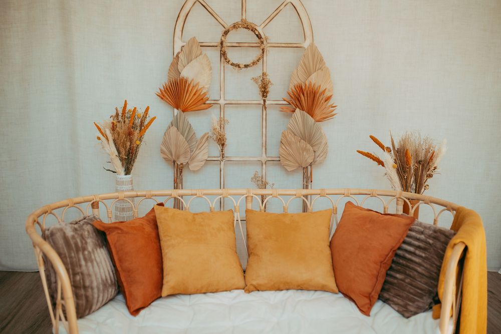 Boho rattan sofa with orange and brown pillows, dried pampas grass decor, and woven wall art on a light background.
