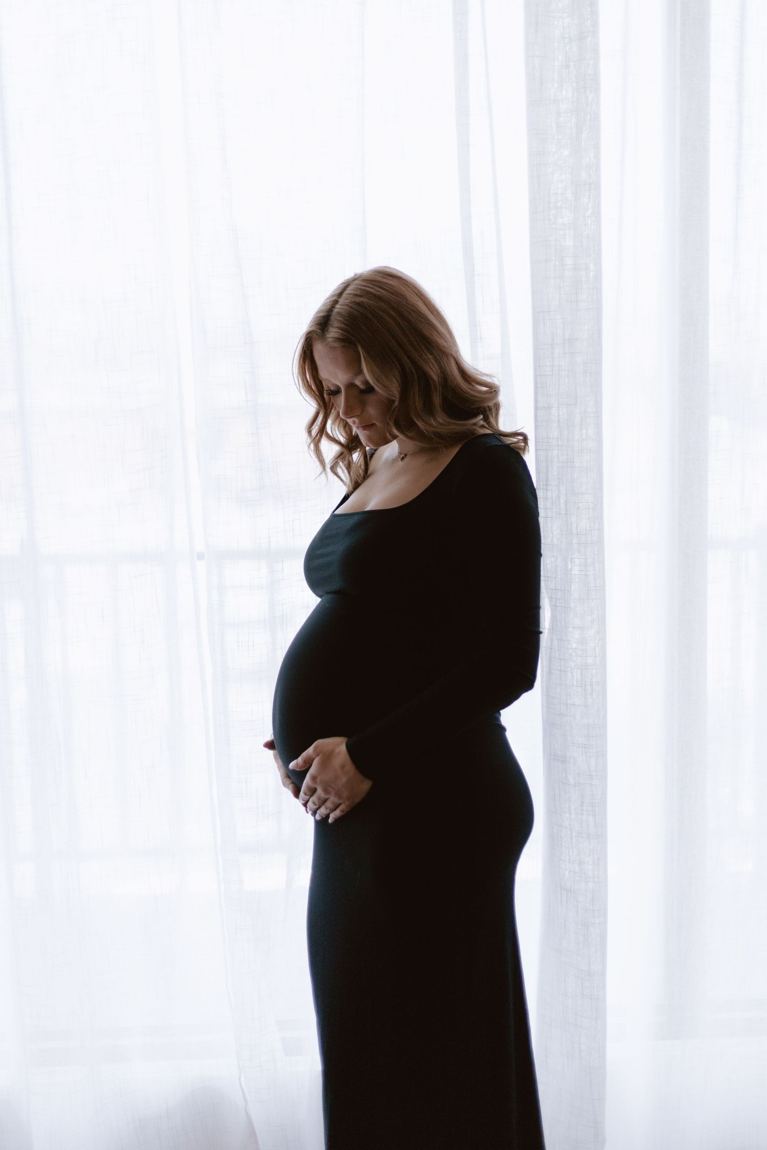 Pregnant woman in black dress cradling baby bump by sheer white curtains.