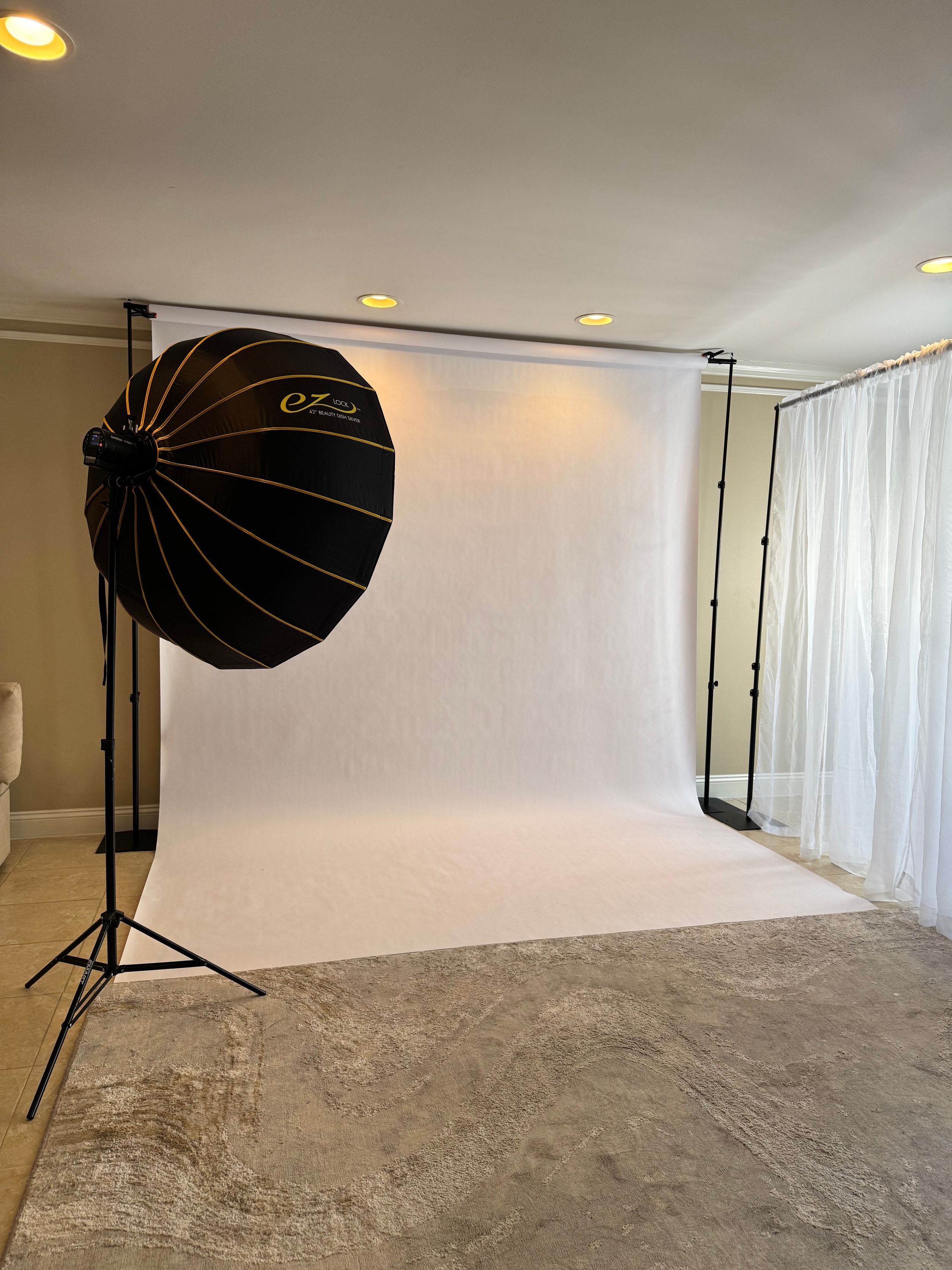 Professional photography studio setup with large parabolic softbox light, white backdrop, and light stand on carpeted floor.