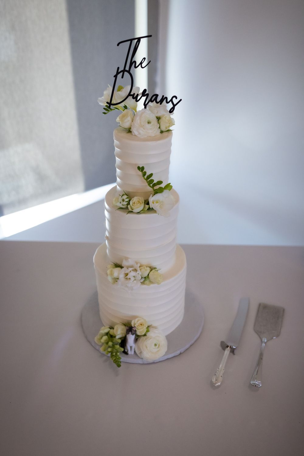 Three-tier white wedding cake with roses and The Durans cake topper on a table.
