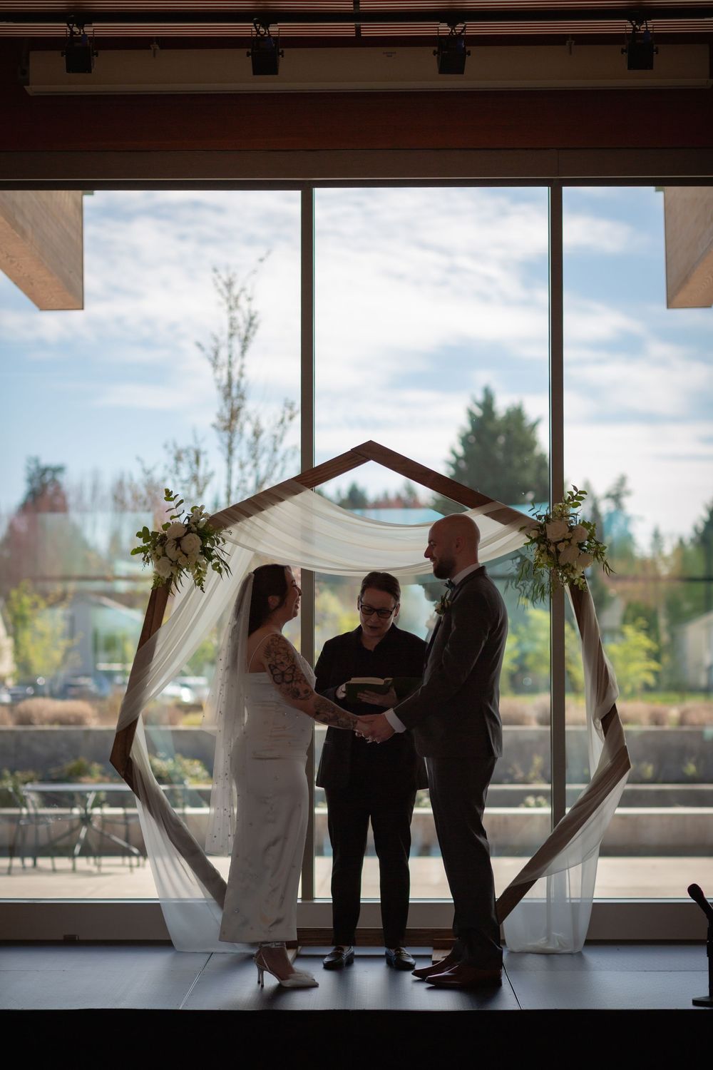 Bride and groom exchanging vows under hexagonal arch with officiant at indoor wedding.