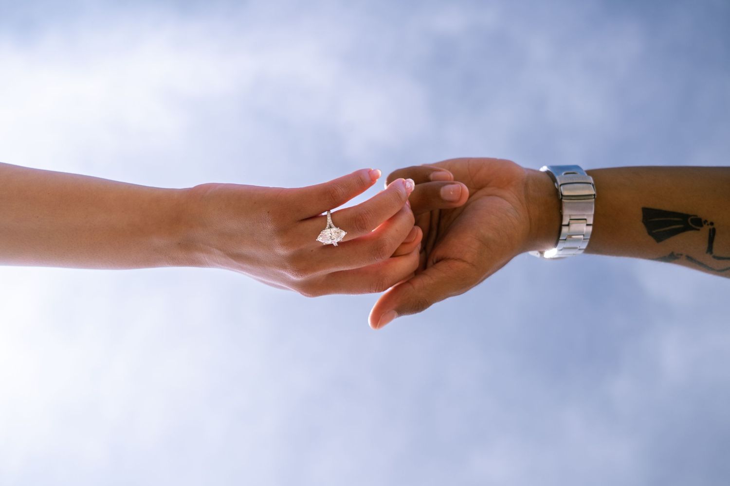 Two hands touching with an engagement ring and a watch against a blue sky backdrop. Carlsbad, CA