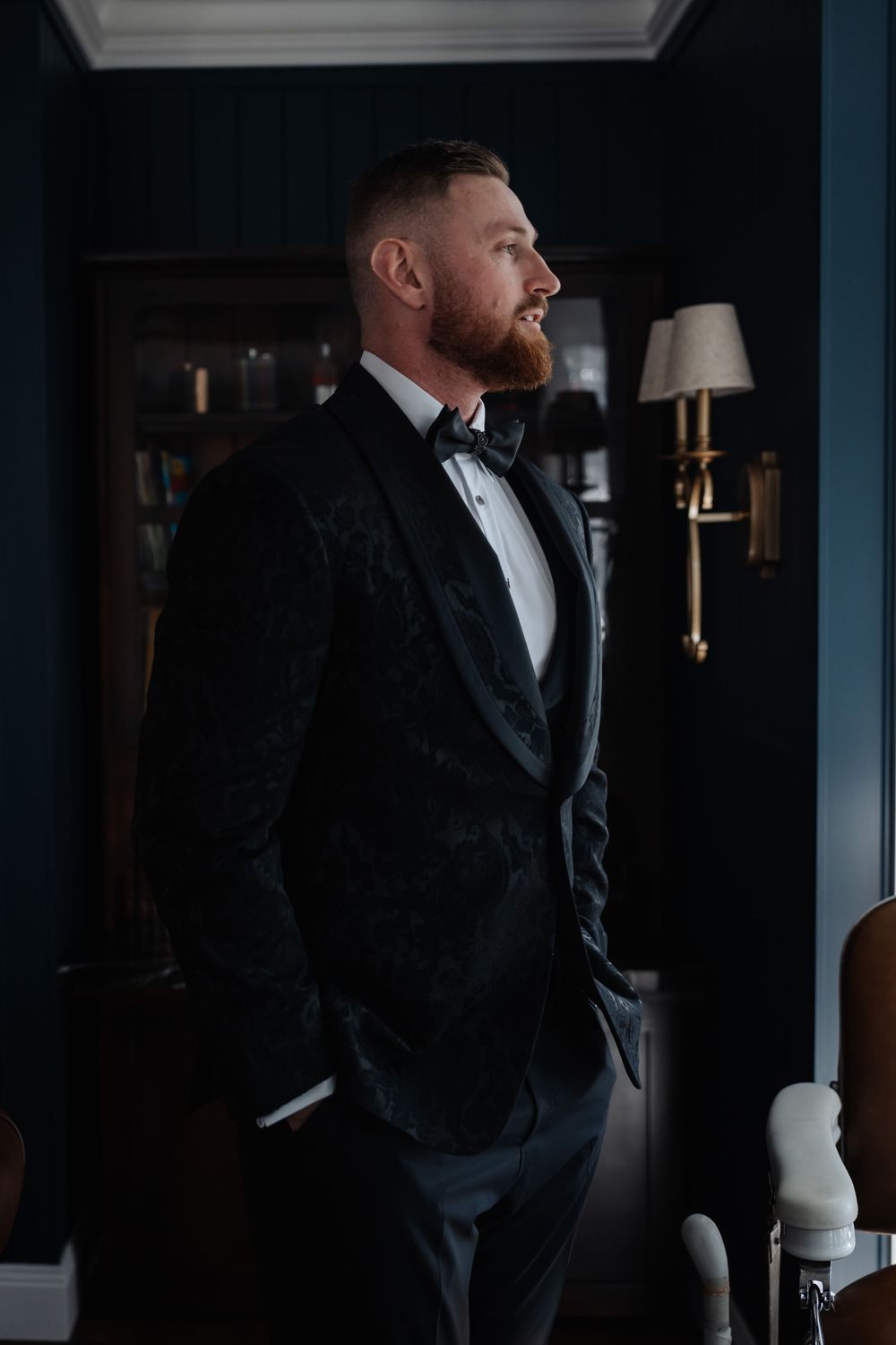 A bearded groom in a black velvet tuxedo with bow tie stands confidently in an elegant dark-walled room.