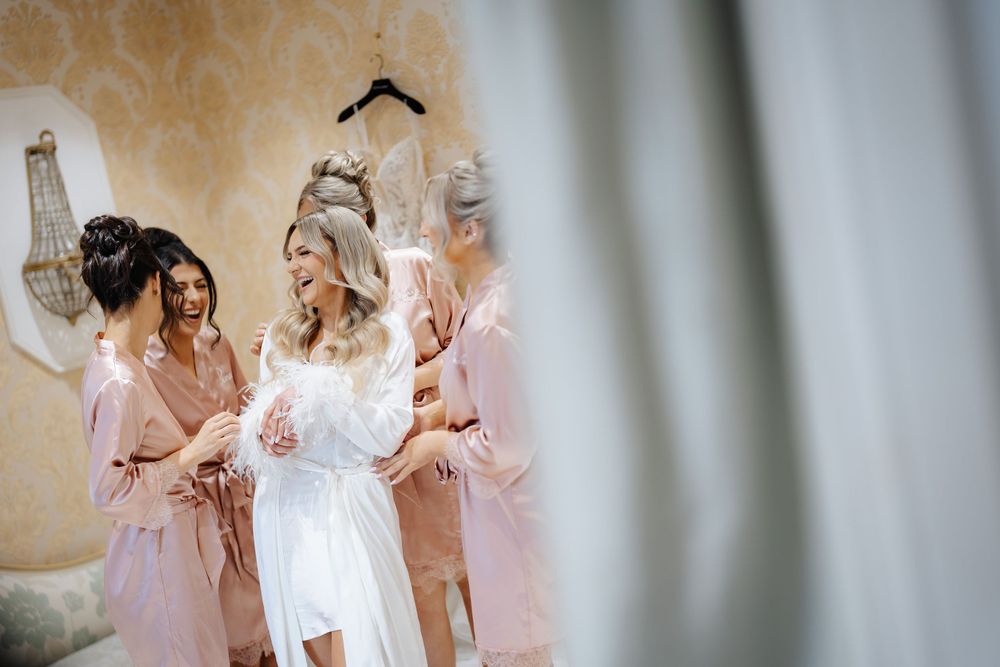 Bride in white robe laughing with bridesmaids in pink robes during wedding morning preparations.