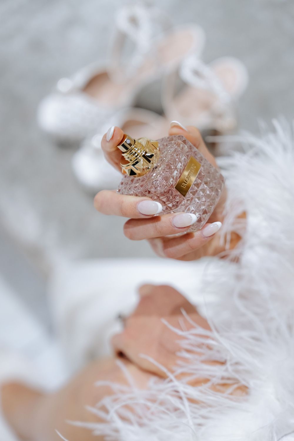 A bride holds a crystal perfume bottle with gold cap, wearing white feather robe, with bridal heels in background.