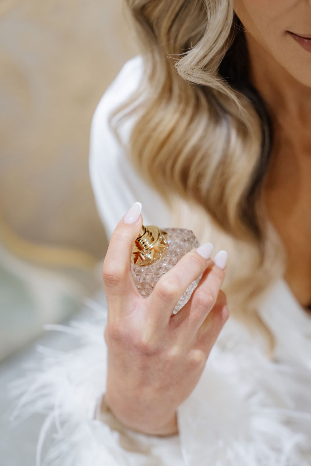 Blonde woman in white robe holding a crystal perfume bottle with a gold ring on her finger.