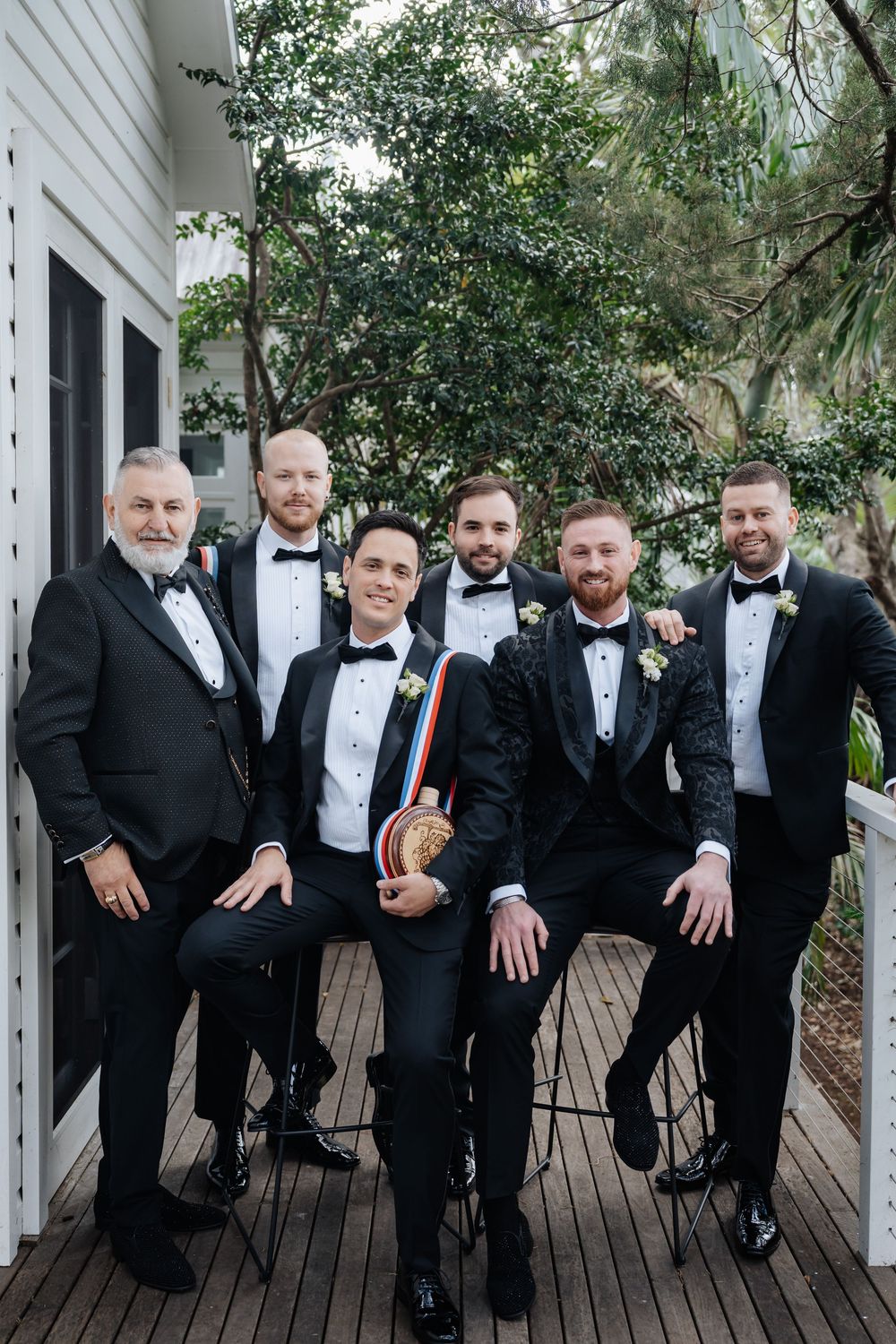 Six groomsmen in black tuxedos pose on a deck, with the groom holding a decorative sash at a wedding.