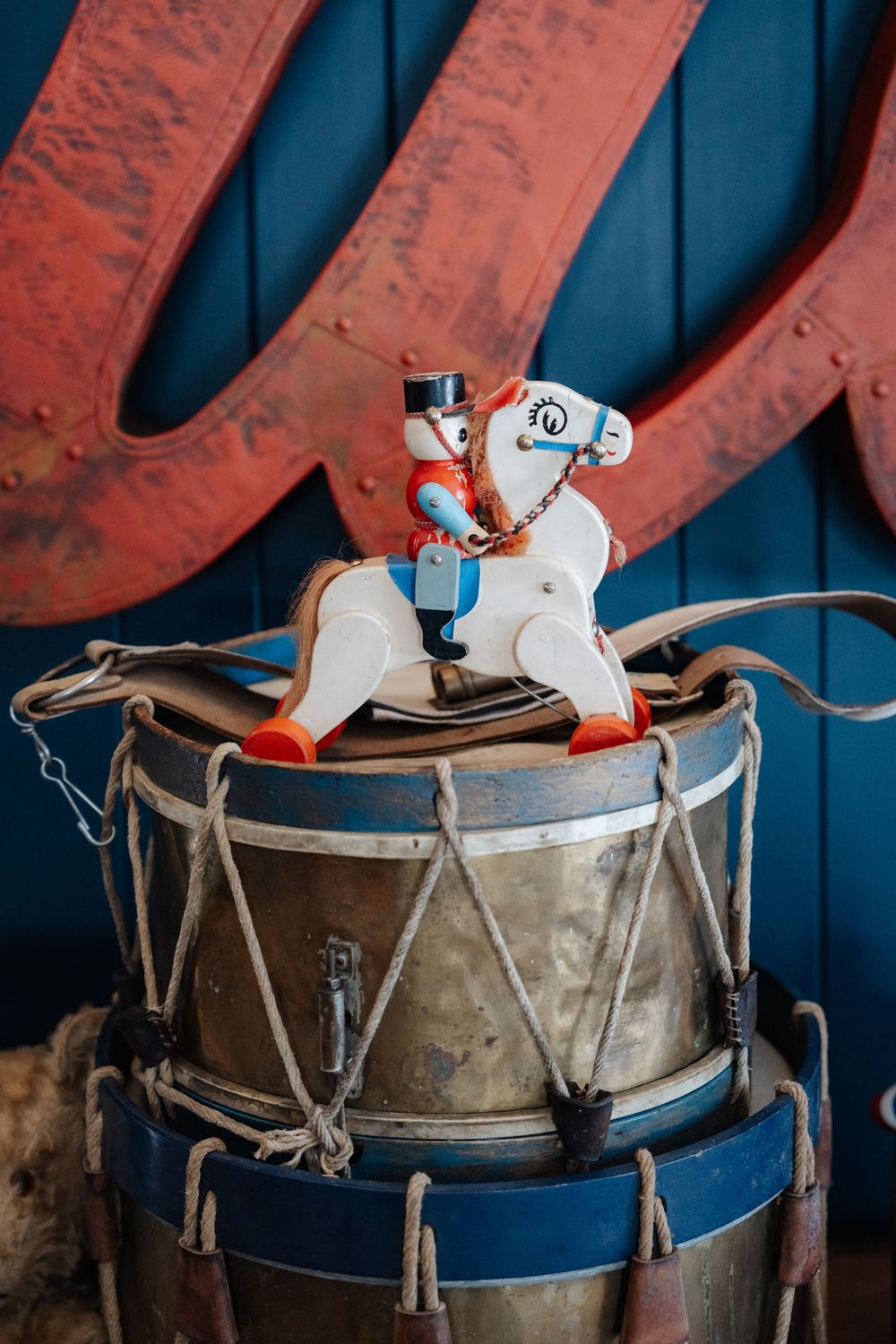 Vintage toy soldier riding a rocking horse atop antique drums, displayed against a blue wall with red metal letters.