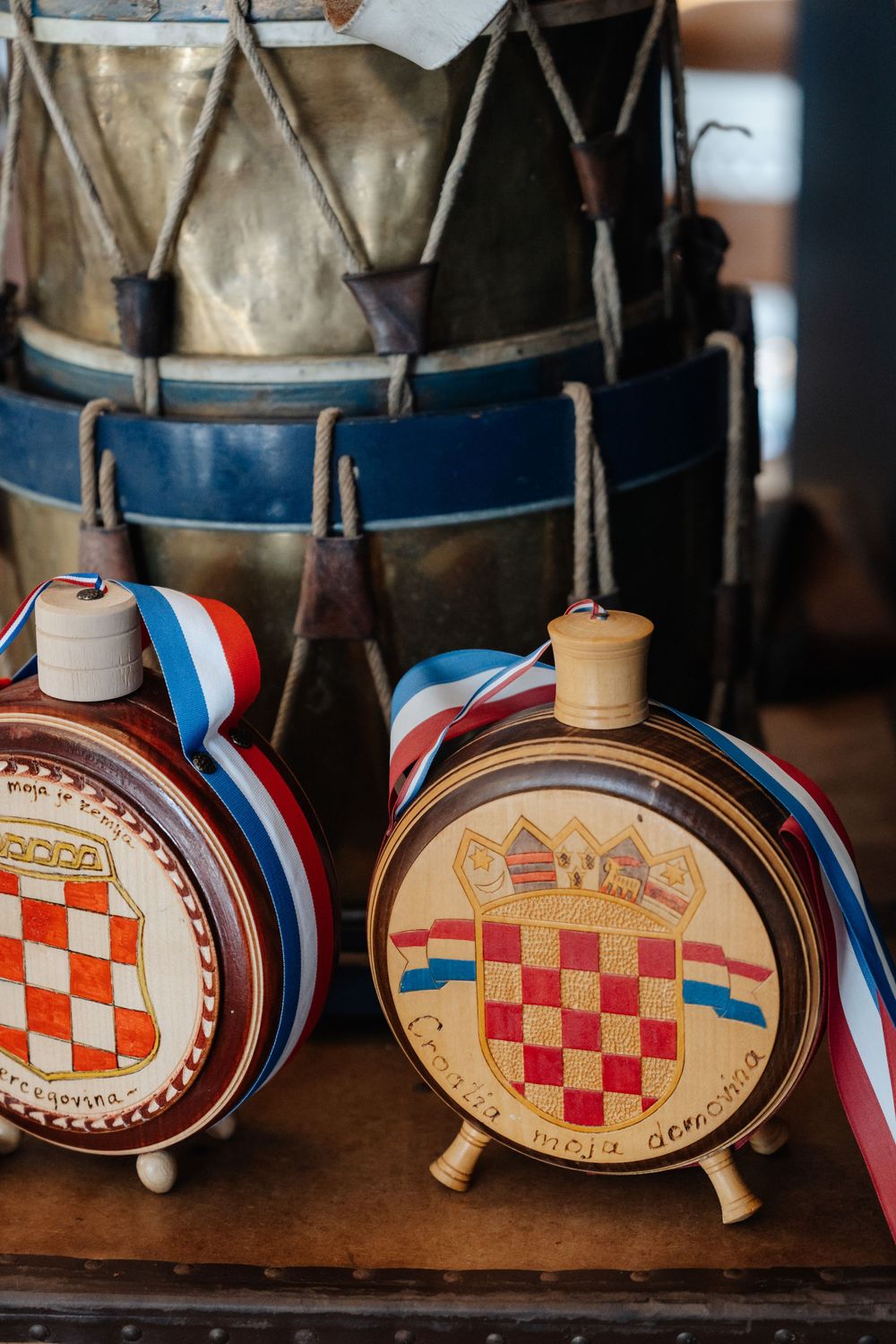 Two wooden Croatian coat of arms canteens with red-white-blue ribbons displayed in front of vintage drums.