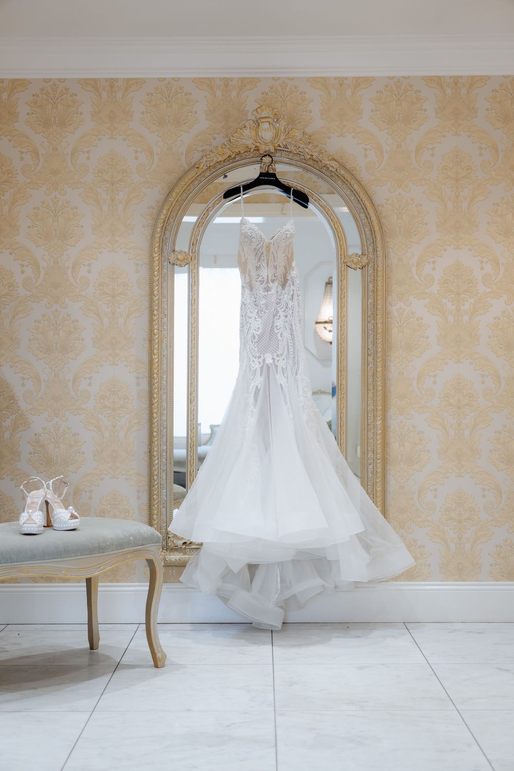 White lace wedding dress hanging on an ornate gold mirror in an elegant bridal suite with damask wallpaper.