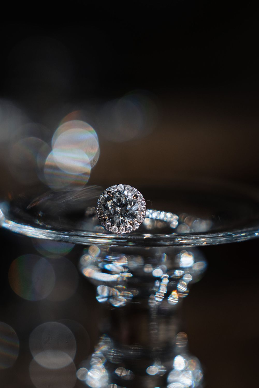 Elegant diamond halo engagement ring displayed on glass surface with bokeh light reflections on dark background.