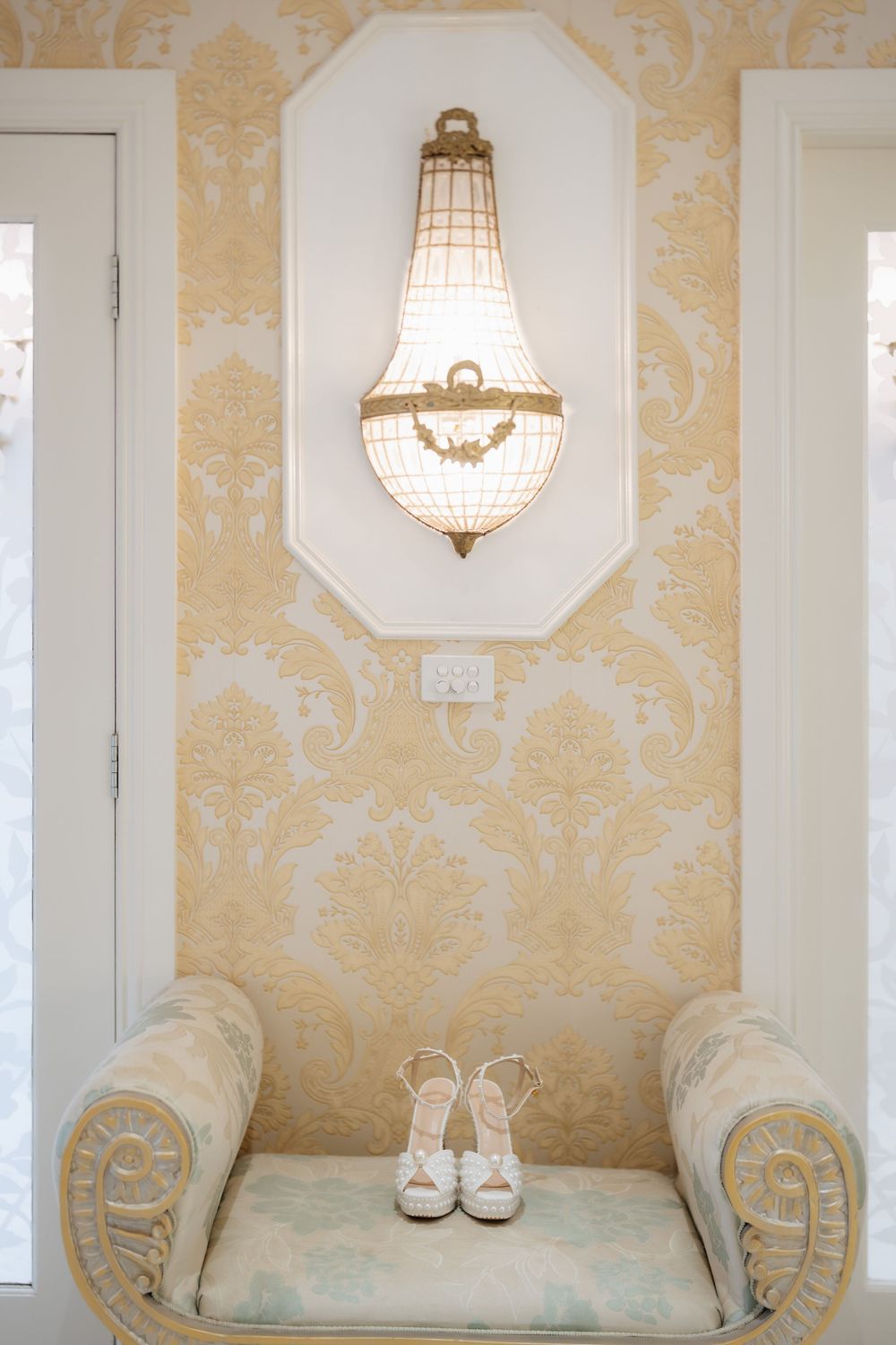 Elegant gold damask wallpaper with ornate wall sconce above a vintage bench holding white bridal heels.