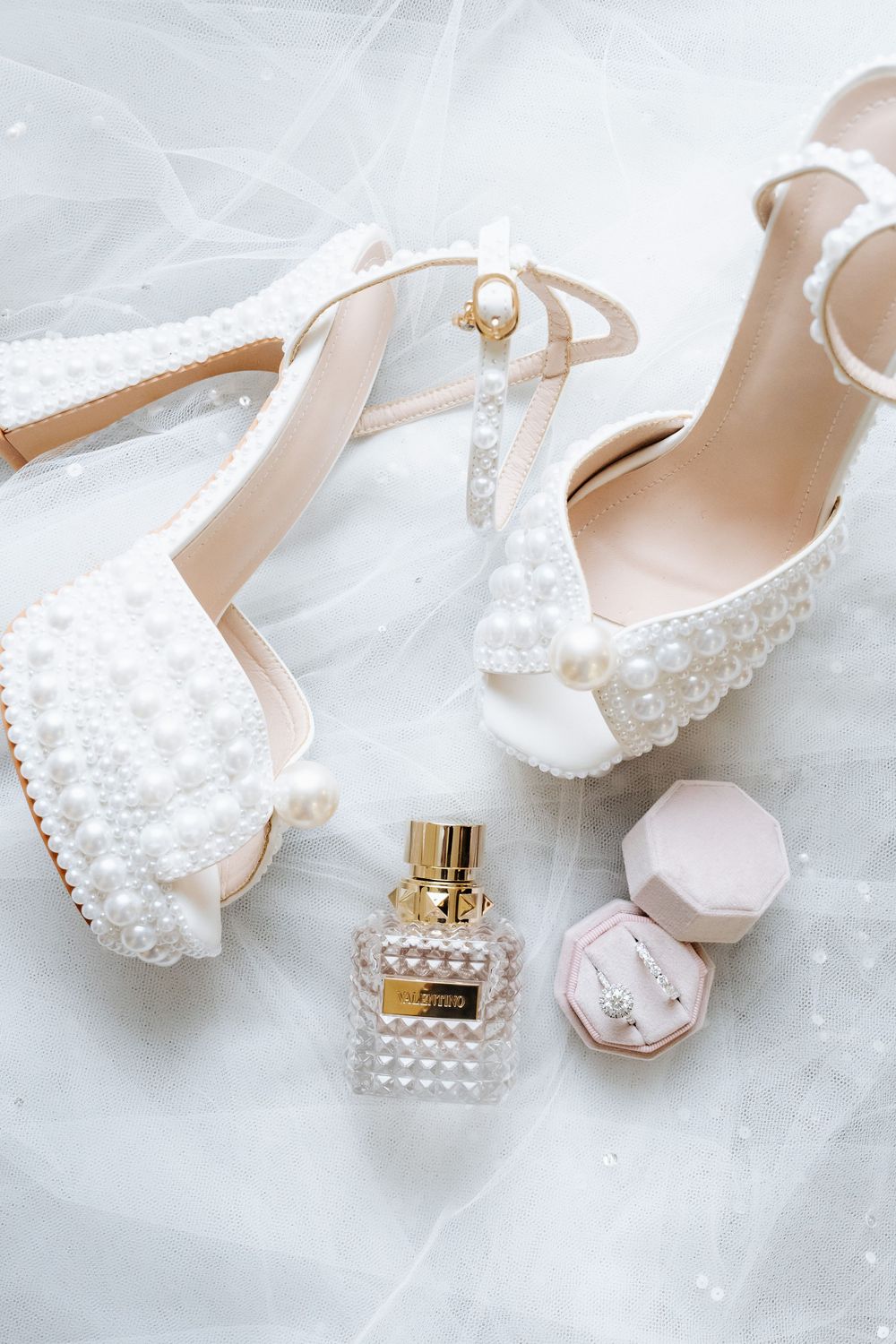 Pearl-embellished bridal heels displayed with a perfume bottle, pink jewelry box, and delicate necklace on white veil fabric.
