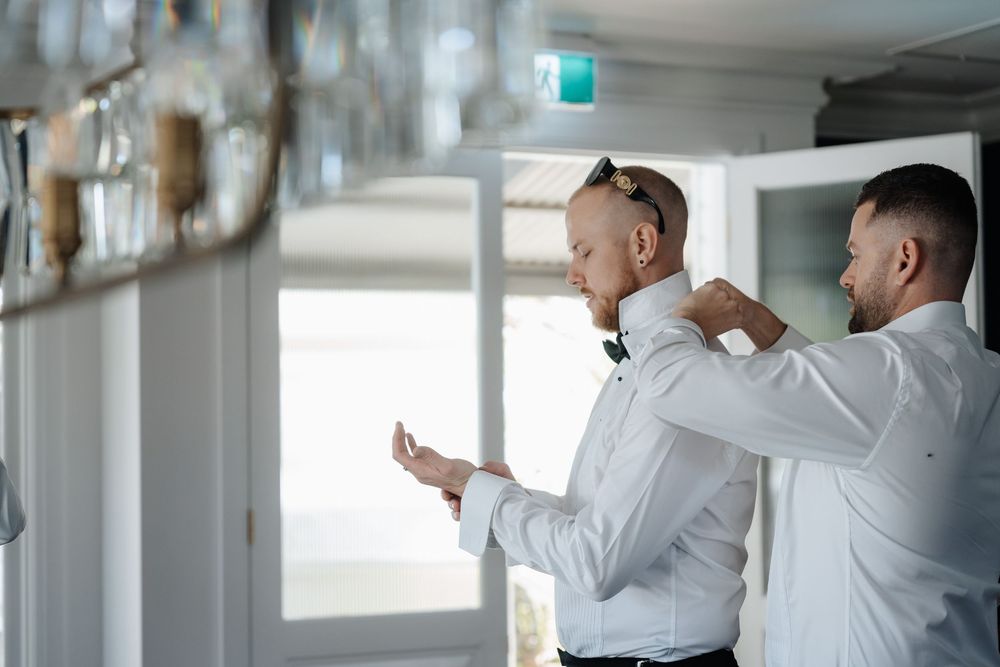 Groomsmen helping groom adjust green bow tie and cufflinks before wedding ceremony indoors.