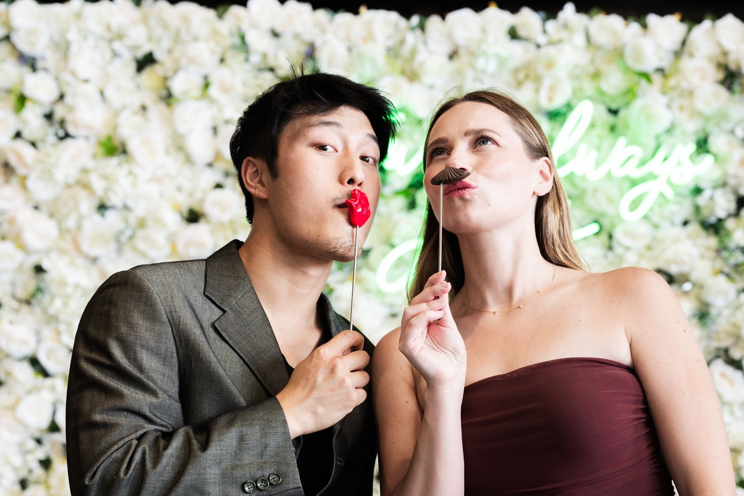 Young couple posing playfully with red lips props in front of a white floral photo booth backdrop.