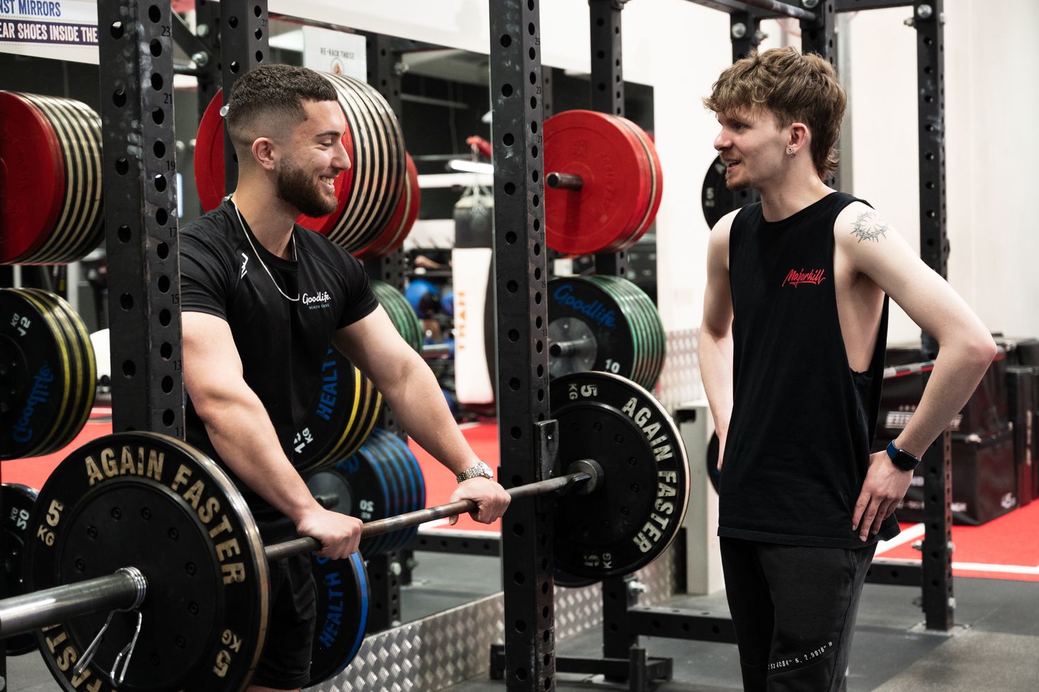 Two young men talking in a gym surrounded by barbells and weight racks during a training session.