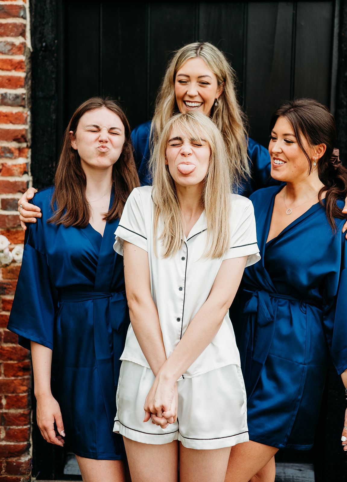 Four women in satin robes and pajamas posing playfully, making funny faces together in a bridal party getting-ready photo.