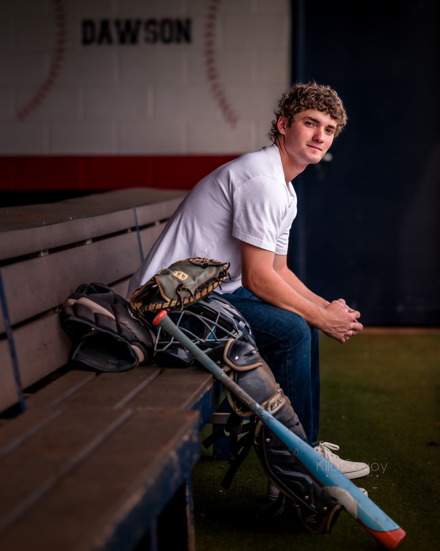 Young baseball player sits in dugout with glove, helmet, and bat, wearing white tee and jeans at Dawson field.