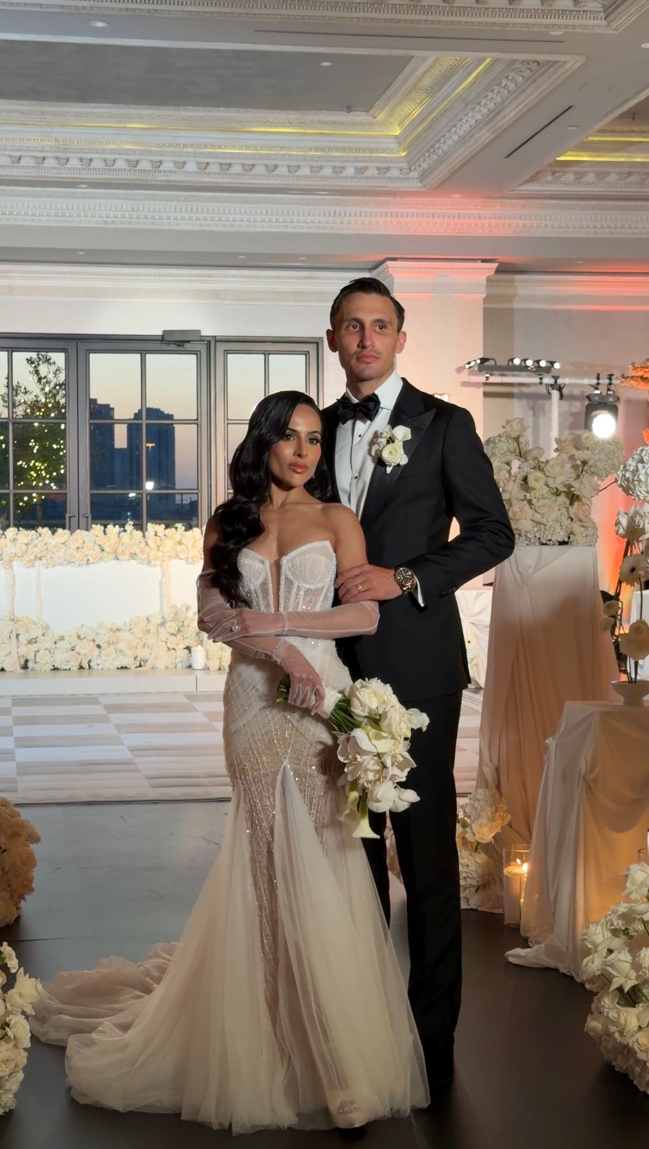 Elegant bride in mermaid gown holding white bouquet stands with groom in black tuxedo at luxurious wedding reception.