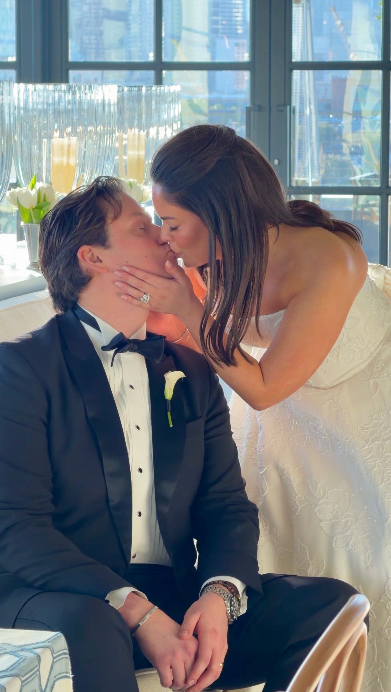 A bride and groom share a romantic kiss at their wedding reception, with the groom in a black tuxedo and bride in white.