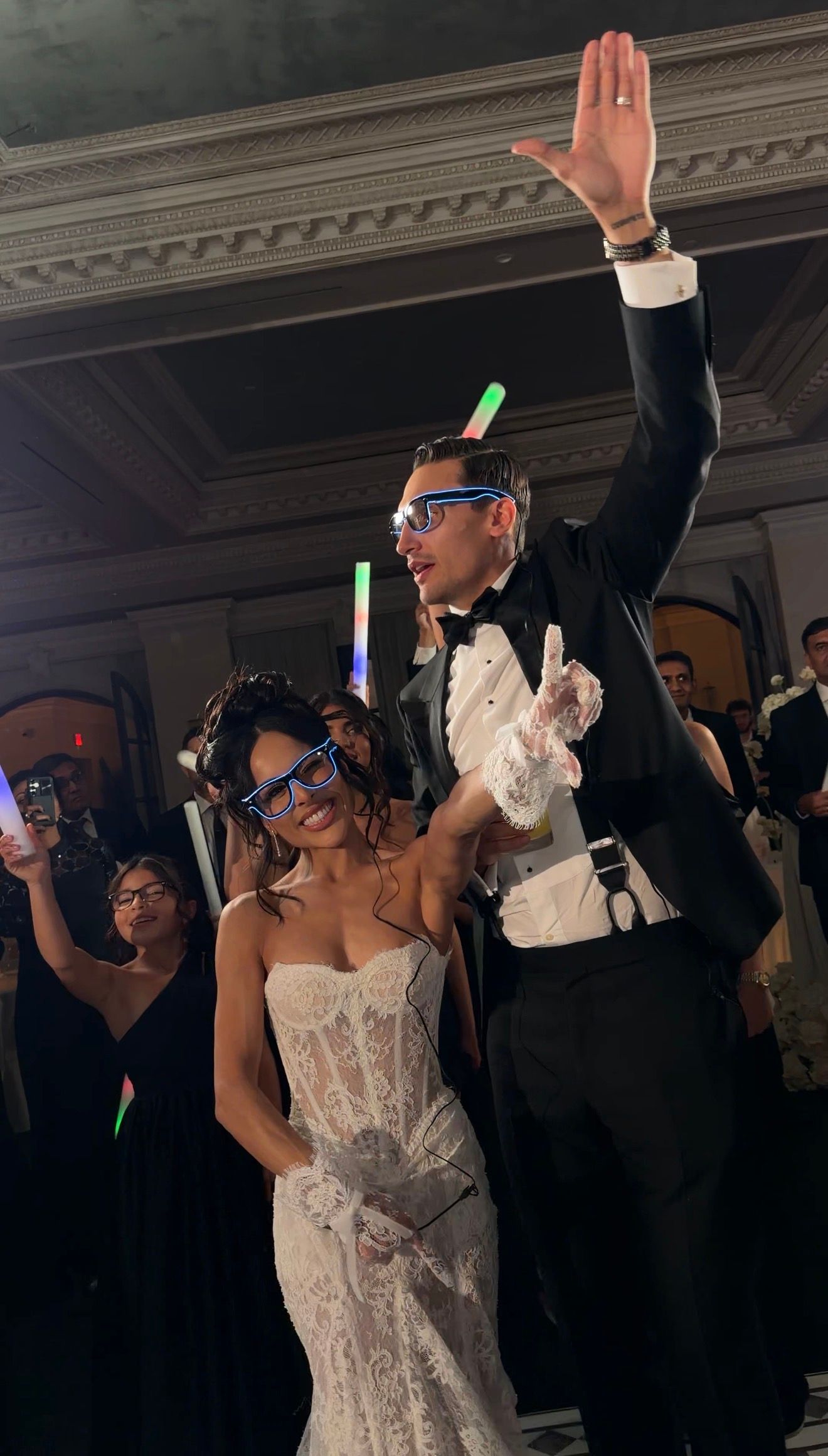 Bride in lace gown and groom in tuxedo celebrate at wedding reception with glow sticks and LED glasses.
