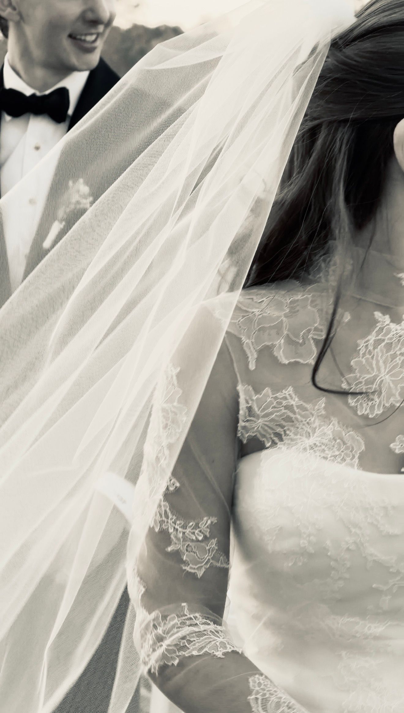 Black and white photo of a bride in lace wedding dress and veil standing beside a groom in a tuxedo.