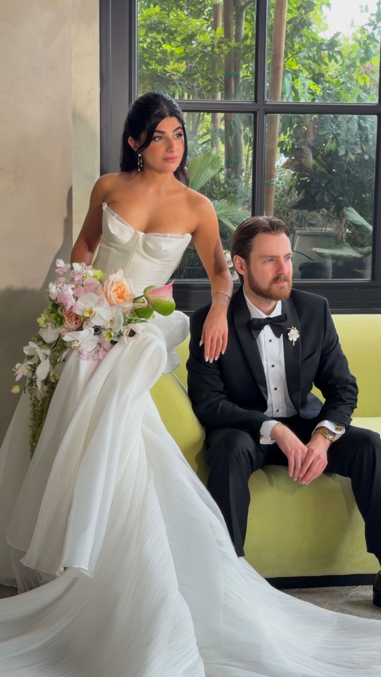 Elegant bride in strapless white gown holding floral bouquet poses with groom in black tuxedo on yellow sofa.