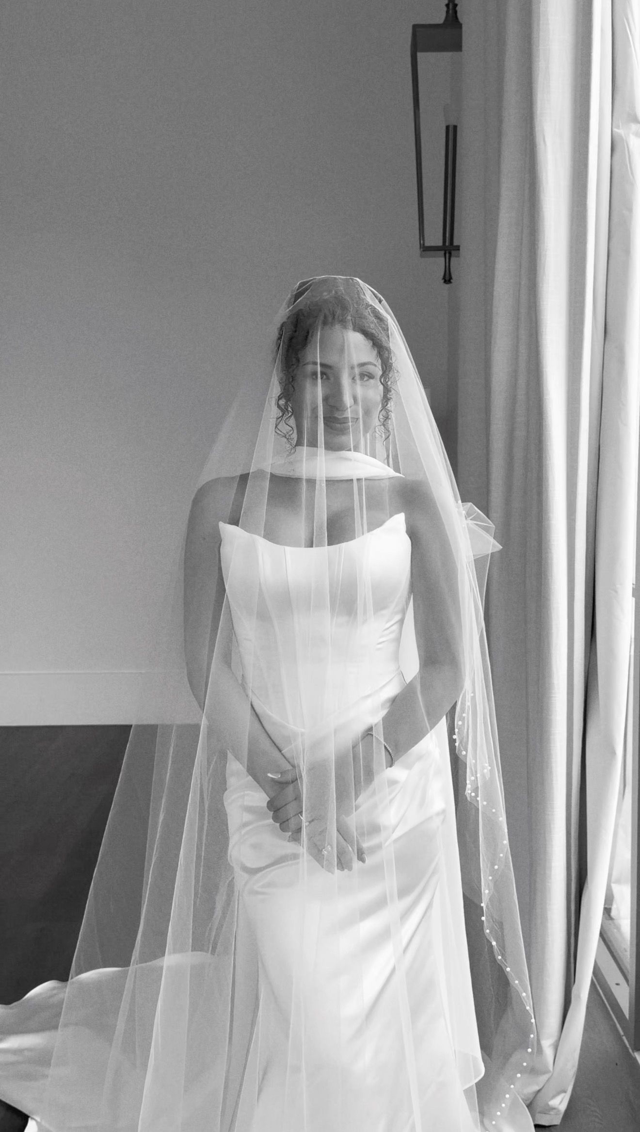 Bride in strapless white wedding gown and long veil poses elegantly in black and white photo near window.