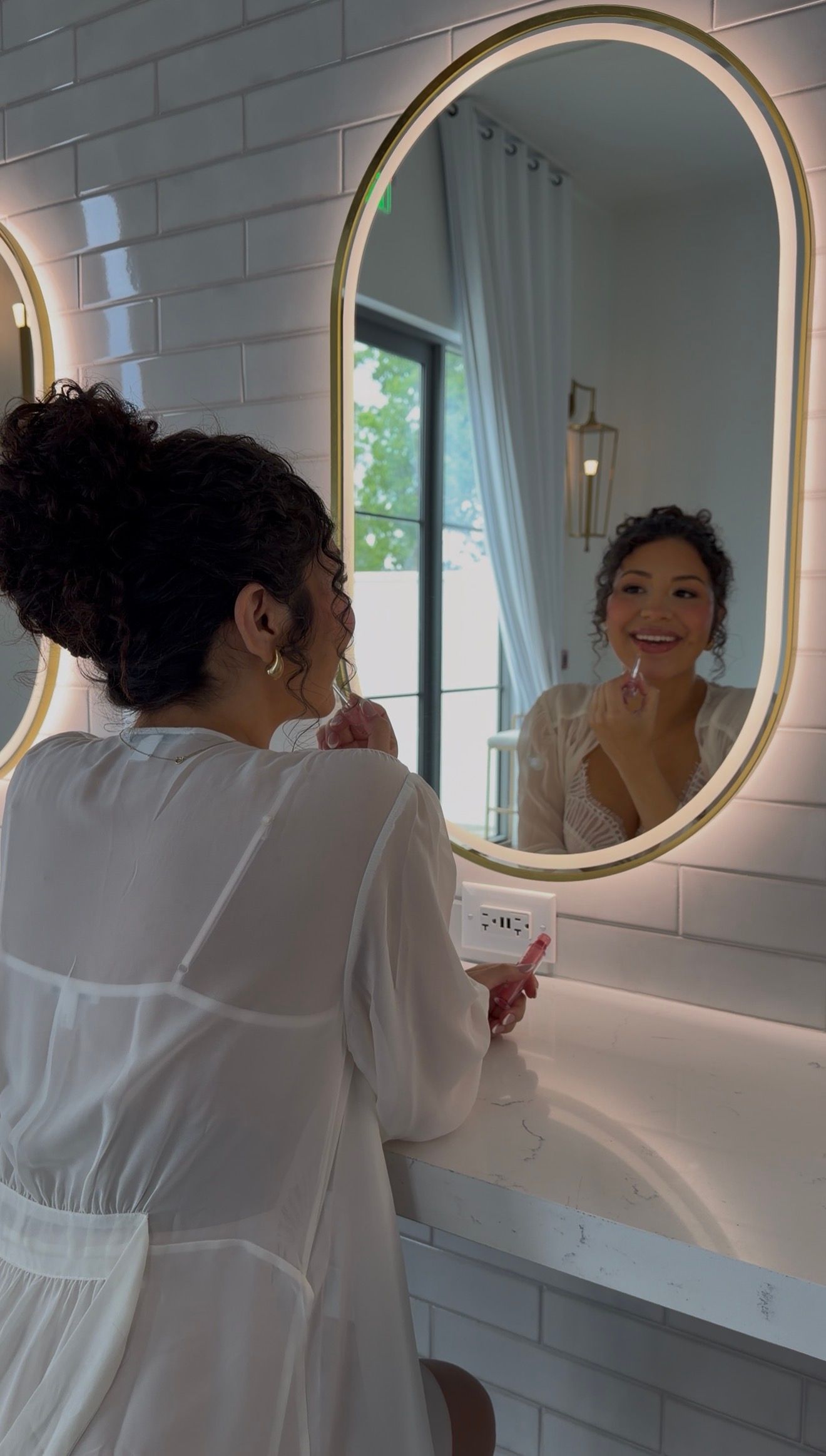 Woman in white dress applying lipstick while looking into an illuminated oval gold-framed bathroom mirror.