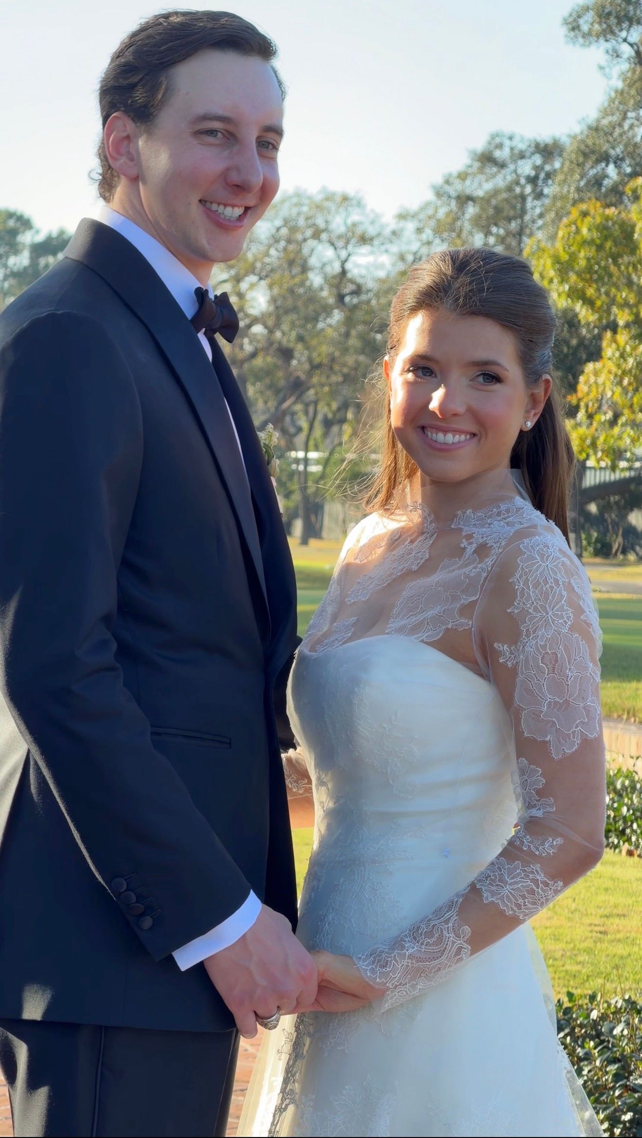 Happy bride and groom posing outdoors, she in lace wedding gown, he in dark suit on wedding day.