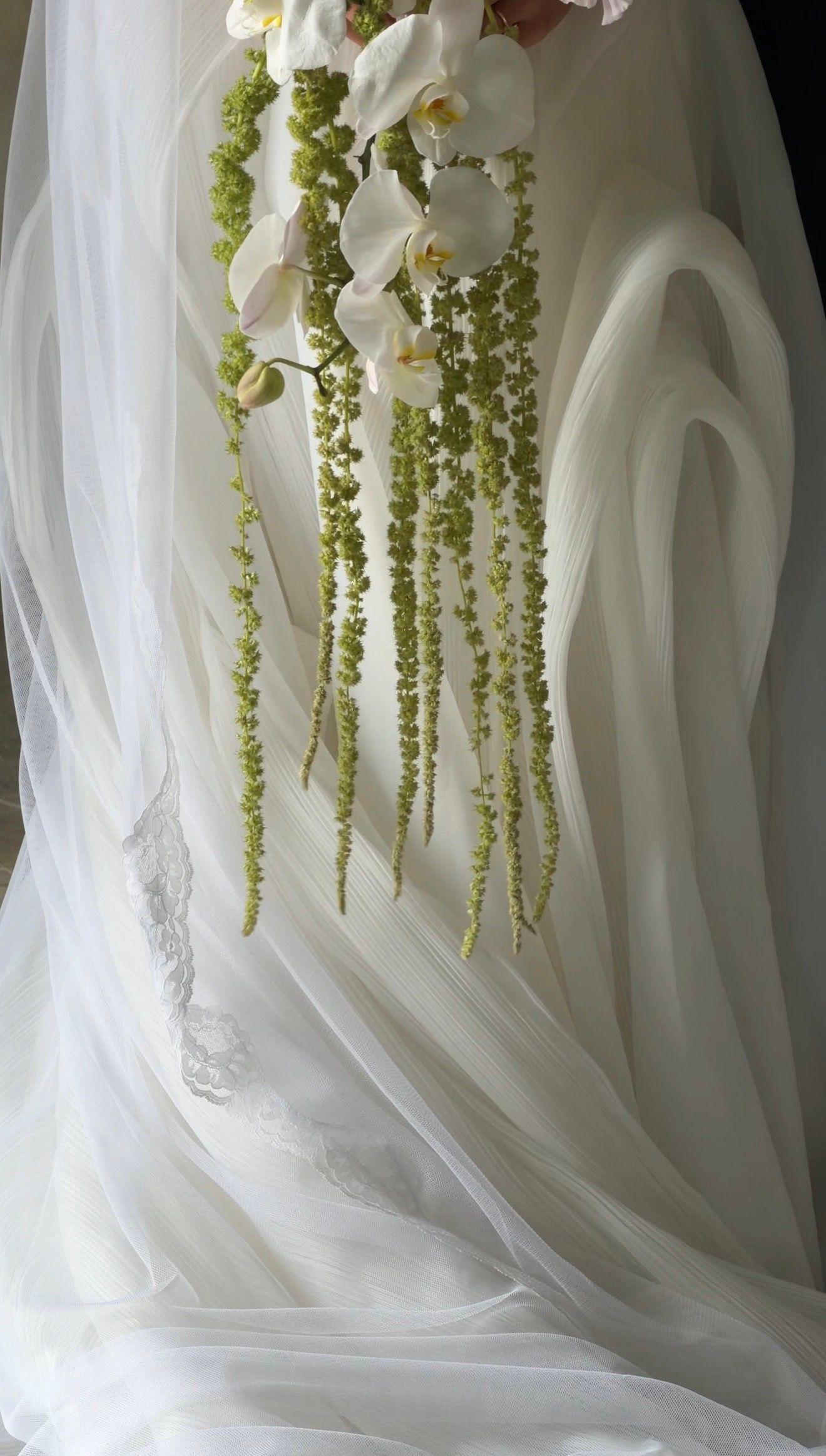 Elegant bridal bouquet with white orchids and cascading green amaranthus against a flowing white veil.