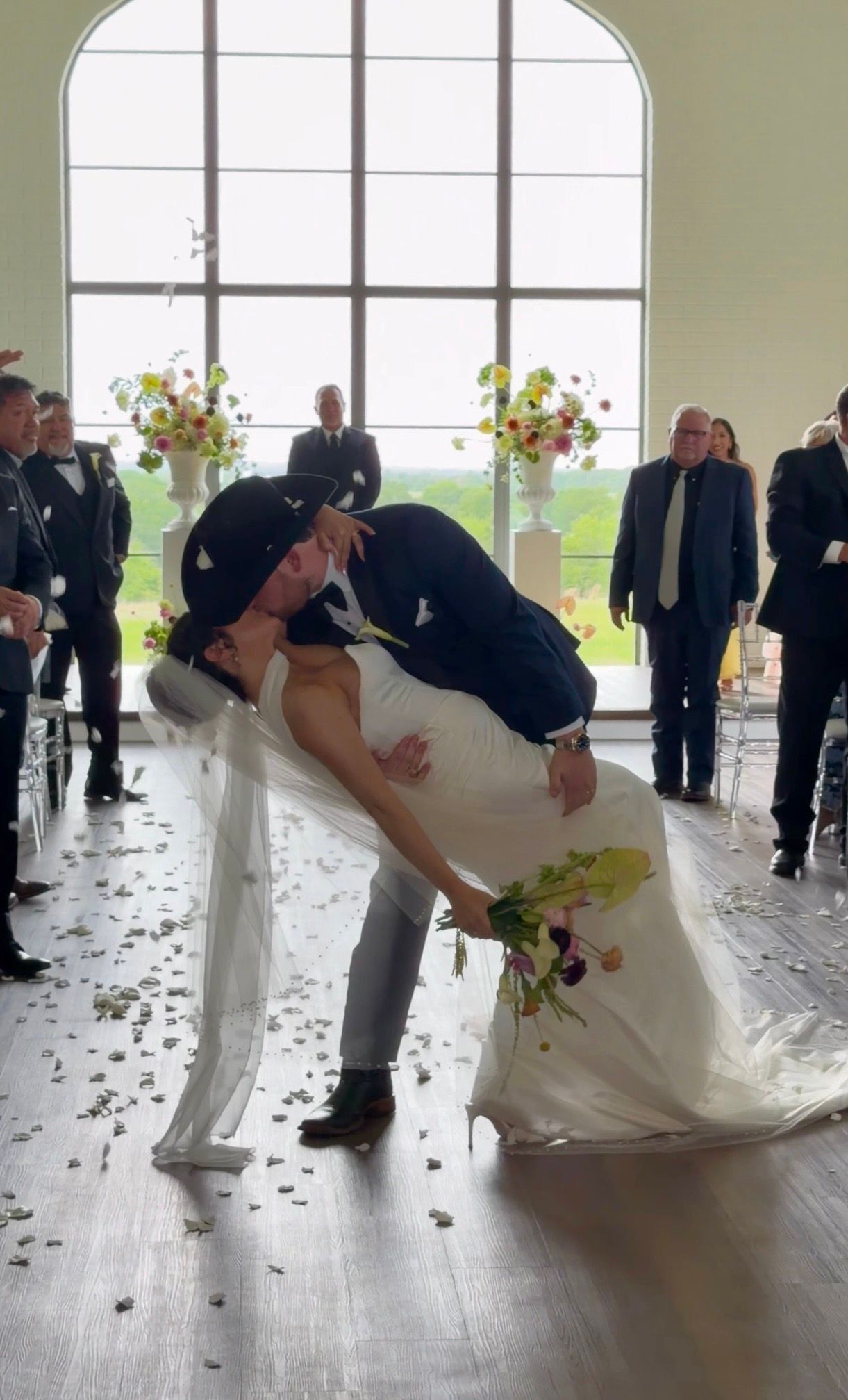 Bride and groom share a romantic dip kiss at wedding ceremony with floral arrangements and guests in background.