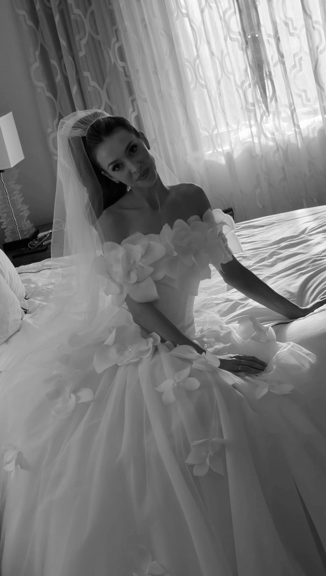 Black and white photo of a bride in an elegant off-shoulder ball gown with floral details and veil, seated on a bed.