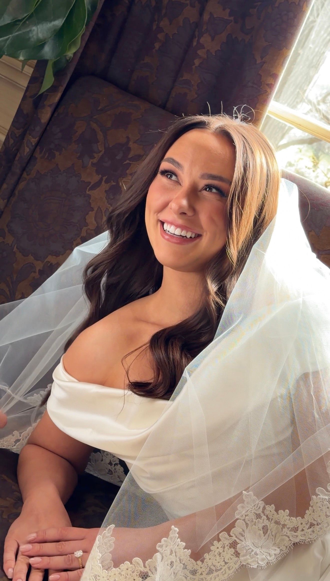 Smiling bride in white off-shoulder gown and veil with wavy brunette hair on her wedding day.